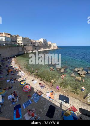 Tourists sunbathing and swimming in the clear waters of the Mediterranean Sea below the historic waterfront of Ortigia in Siracusa, Sicily, Italy Stock Photo