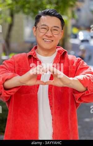 I love you. Asian middle-aged man makes symbol of love, showing heart sign to camera express romantic feelings, express sincere positive feelings outdoors. Charity, gratitude, donation. on city street Stock Photo