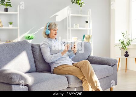 Senior woman at home listening to music, using smartphone and big headphones set, looking at phone Stock Photo