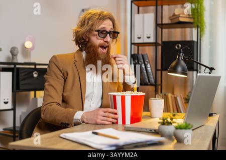 Caucasian businessman freelancer taking break from work wearing 3D glasses earing popcorn and watching movie on laptop. Smiling bearded man guy sitting at home office desk having snacks during break Stock Photo