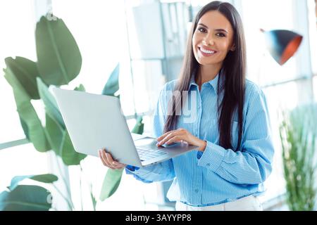 Confident young businesswoman working on a laptop in a stylish office setting with greenery, showcasing professionalism Stock Photo