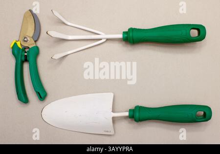Three white and green gardening tools - a trowel, a hand rake, and a pruner - on a light grey/tan background. Stock Photo