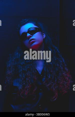 Fashion portrait of young woman with curly hair and sunglasses standing under blue and red neon glow against gradient background. Stock Photo