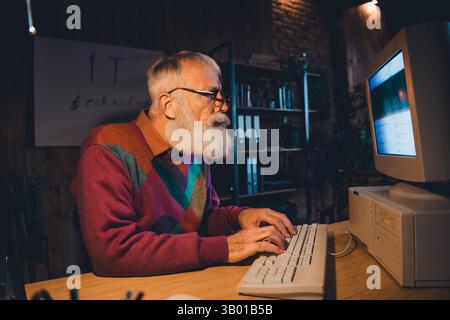 Elderly man coding on a vintage computer in a cozy office with soft evening light, showcasing technology and determination Stock Photo