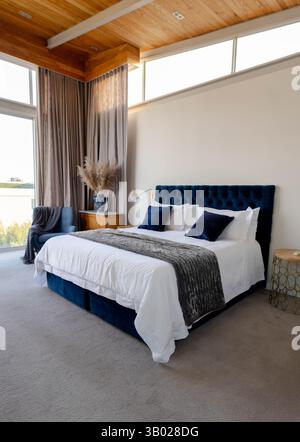King-size bed sitting in modern bedroom, featuring navy headboard, white linens and pampas grass Stock Photo