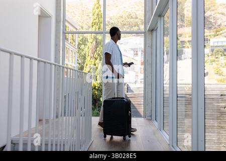 African American man pulling suitcase and checking smartphone in modern corridor with glass windows Stock Photo