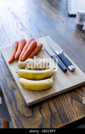 Wooden cutting board is displaying bananas, ginger root, carrots and knives on dining table Stock Photo