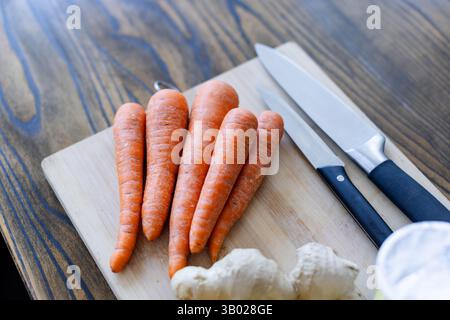 Light cutting board is resting on kitchen table, displaying carrots, knives and ginger root Stock Photo