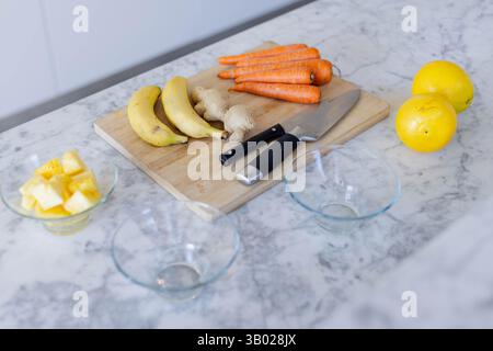 Wooden cutting board featuring ginger, carrots on marble countertop with pineapple bowl and knives Stock Photo