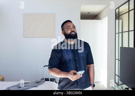 Holding smartphone African American man standing in modern bedroom, with suitcase and unmade bed Stock Photo
