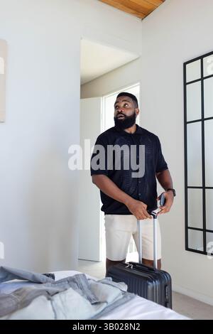 Standing African American man holding smartphone in minimalist bedroom, with black rolling suitcase Stock Photo