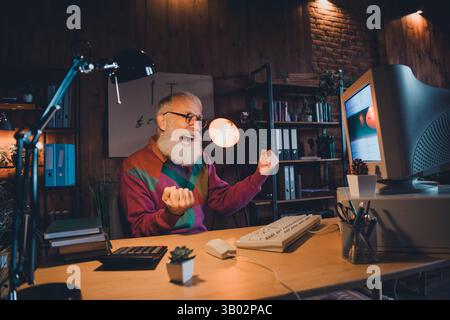 Senior man enjoying programming success at home office desk with vintage computer setup in cozy ambient light Stock Photo