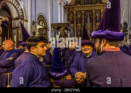 Two brotherhood members in purple robes and pointed hoods shake hands with a smile as the brotherhoods prepare for the Good Friday procession Camino del Calvario in the parish church of El Salvador. Plaza Salvador, Cuenca, Castilla-La Mancha, Spain. During Semana Santa in Cuenca, 33 Hermandades take part in eleven processions through the city during Holy Week. Stock Photo