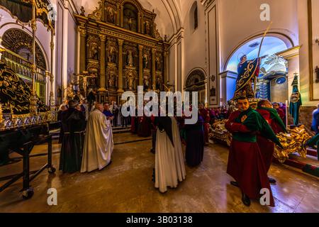 Clergy and brotherhood members gather in front of the main altar of the parish church of El Salvador to prepare for the Good Friday procession Camino del Calvario. Plaza Salvador, Cuenca, Castilla-La Mancha, Spain. On the right, a member in red and green robes carries a golden processional staff, while a statue of Christ from the procession can be seen in the background. During Semana Santa in Cuenca, 33 Hermandades take part in eleven processions through the city during Holy Week. Stock Photo