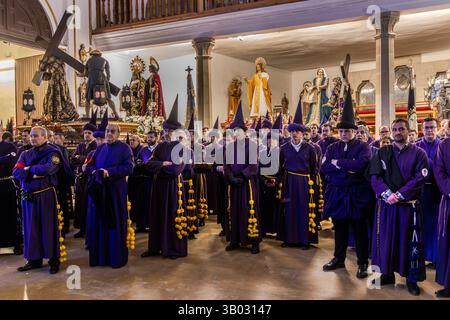 Numerous brotherhood members in purple robes and pointed hoods with yellow pompom tassels wait packed together in the parish church of El Salvador ahead of the Good Friday procession Camino del Calvario. Plaza Salvador, Cuenca, Castilla-La Mancha, Spain. Above them, processional statues already stand on a gallery, including a figure of Christ carrying the cross. During Semana Santa in Cuenca, 33 Hermandades take part in eleven processions through the city during Holy Week. Stock Photo
