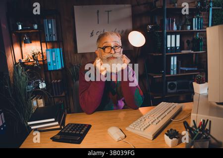 Elderly programmer working at desk in casual sweater in cozy home office setting under warm evening light Stock Photo