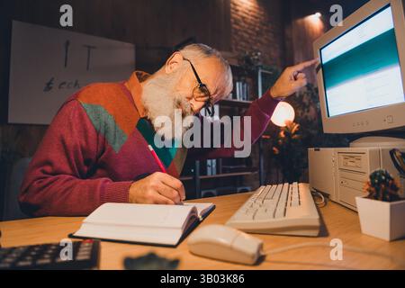 Elderly programmer working attentively at his desk in cozy evening light, taking notes and analyzing data on desktop computer Stock Photo