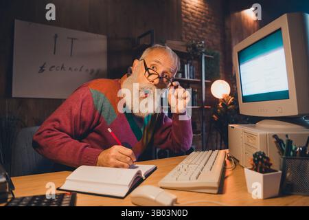 Elderly man working on a computer in cozy office setting with intense expression and colorful sweater, late evening atmosphere. Stock Photo