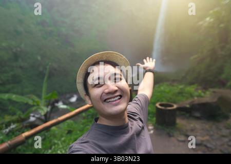 Young Asian man with straw hat taking selfie portrait with smile face expression while showing the waterfall Stock Photo
