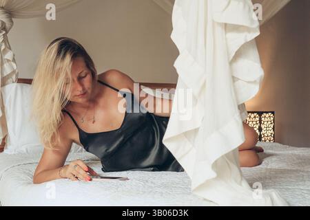 Beautiful blonde woman using smartphone lying on bed in bedroom Stock Photo