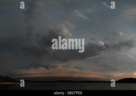 Powerful and dramatic scene of dark storm clouds sweeping over a still lake just before nightfall. The heavy clouds are highlighted by a moody sunset Stock Photo