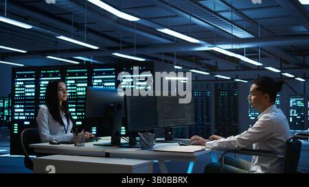 Person with paraplegia helped by coworker in data center to arrive at desk, coding on PC. Woman in wheelchair doing maintenance on server room infrastructure rackmounts, camera B Stock Photo