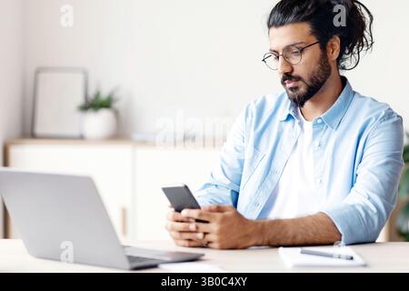 Arab Freelancer Man Messaging On Smartphone, Sitting At Desk In Home Office Stock Photo