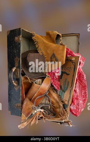 Antique suitcase, handle & latches is open with western gear wear tumbling out. Open antique suitcase stands on end with old cowboy western clothes. Stock Photo