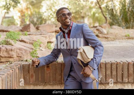 Elegant businessman wearing sunglasses with a shoulder bag is leaning on a low wall in a park, smiling and looking away, enjoying his break Stock Photo
