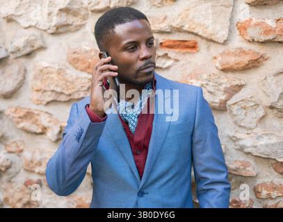 Confident businessman wearing suit and cravat, talking on smartphone while standing in front of old stone wall, looking away with serious expression Stock Photo