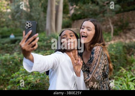 Two multi-ethnic female friends are taking a selfie in a park, sticking out their tongues and making the victory sign with their fingers Stock Photo