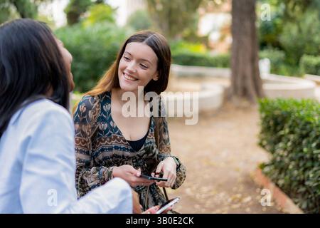 Two young women are using their smartphones and smiling while sitting on a bench in a park, enjoying a moment of relaxation and connection together Stock Photo