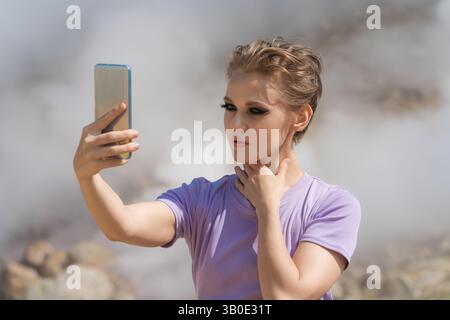 Beautiful female traveler using her cell phone to record self portrait to video call with friends in remote location against backdrop of steaming geys Stock Photo