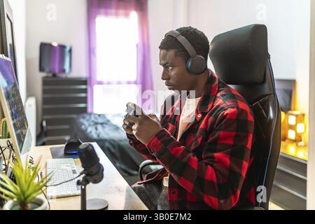 Young man concentrating on playing video games, using headphones and a controller, in his bedroom Stock Photo