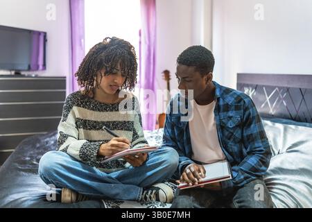 Two focused college students are sitting on a bed, sharing notes and preparing for exams in their dorm room Stock Photo