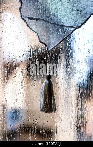 Tassel on net curtain behind rain-splashed window. Stock Photo