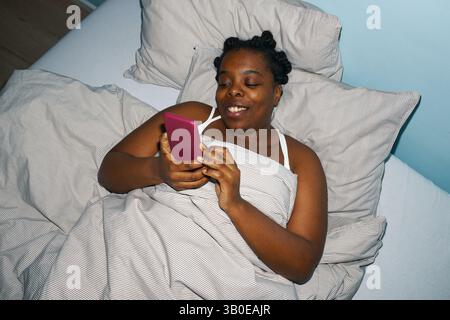 Smiling Black woman lying in bed while holding and using smartphone, with pillows and bedding surrounding her in a cozy setting Stock Photo