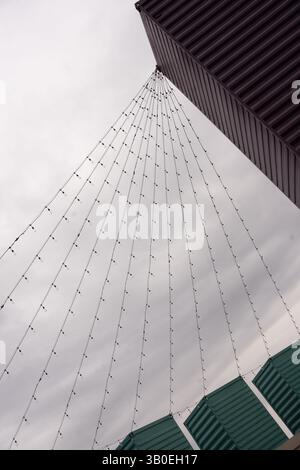 color image  of many strings of light bulbs strung from the tops of three green containers on end to a higher top of a building grey sky background Stock Photo