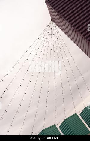 color image  of many strings of light bulbs strung from the tops of three green containers on end to a higher top of a building grey sky background Stock Photo