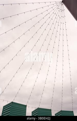black and white of many strings of light bulbs strung from the tops of three containers on end to a higher top of a building creating triangular shape Stock Photo