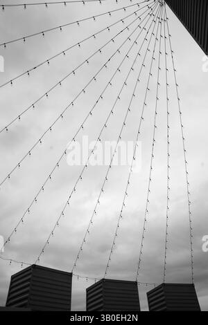 black and white of many strings of light bulbs strung from the tops of three containers on end to a higher top of a building creating triangular shape Stock Photo
