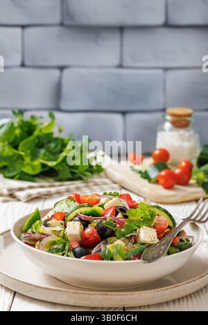 vegetable salad of tomatoes, cucumber, olives, red and green pepper, goat cheese, red onion, cilantro in white bowl on wooden table with brick wall an Stock Photo