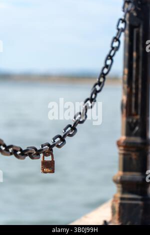 Close-up of a weathered metal chain with a rusty padlock attached, stretching between bollards along a waterfront, with the sea in the background Stock Photo