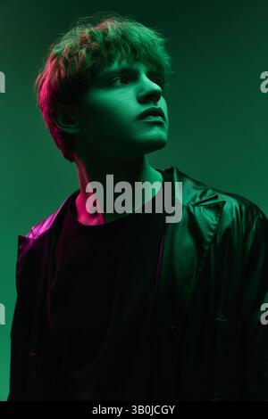 Young man in black outfit and leather coat looks upward with focused gaze under dramatic green neon lighting in minimal studio. Stock Photo