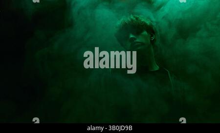 Young man in dark shirt and leather coat stands surrounded by green mist in moody atmosphere against dark studio background Stock Photo