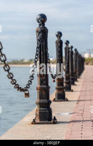 Close-up of a weathered metal chain with a rusty padlock attached, stretching between bollards along a waterfront, with the sea in the background Stock Photo