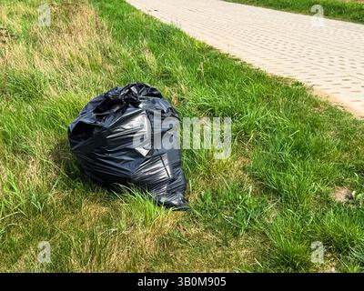 Large black garbage bag placed on green grass near a paved sidewalk, representing outdoor litter or cleanup effort in a park or urban green space duri Stock Photo