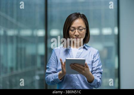 Serious and confident office worker using tablet computer. Asian businesswoman using online financial app, reading thoughtfully while standing near window inside office. Stock Photo