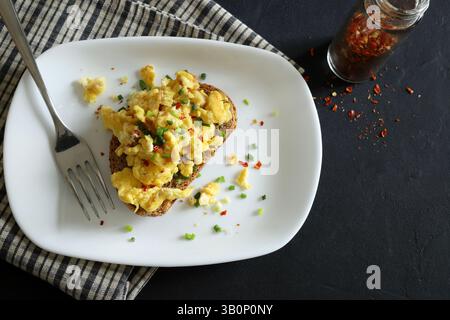 Scrambled eggs with spices and herbs on crunchy whole grain toast on a white plate on a black background. Healthy breakfast. Directly above. Stock Photo