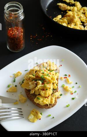 Scrambled eggs with spices and herbs on crunchy whole grain toast on a white plate on a black background. Healthy breakfast. Directly above. Stock Photo
